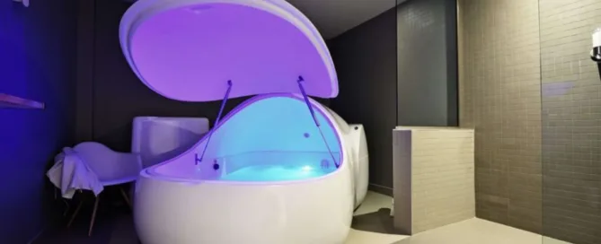 A modern sensory deprivation tank with an open lid, softly illuminated in blue and purple light. It is placed in a dimly lit room with tiled floors and walls. A white chair and towel are visible to the side.