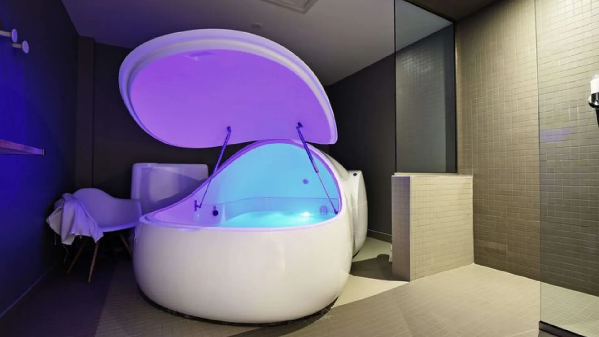 A modern sensory deprivation tank with an open lid, softly illuminated in blue and purple light. It is placed in a dimly lit room with tiled floors and walls. A white chair and towel are visible to the side. A modern sensory deprivation tank with an open lid, softly illuminated in blue and purple light. It is placed in a dimly lit room with tiled floors and walls. A white chair and towel are visible to the side.