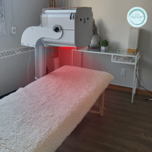 A treatment room with a soft, fleece-covered therapy bed and a large light therapy machine emitting a red glow. A glass table with plants and decor sits nearby, and a SoulTuary logo is on the wall.