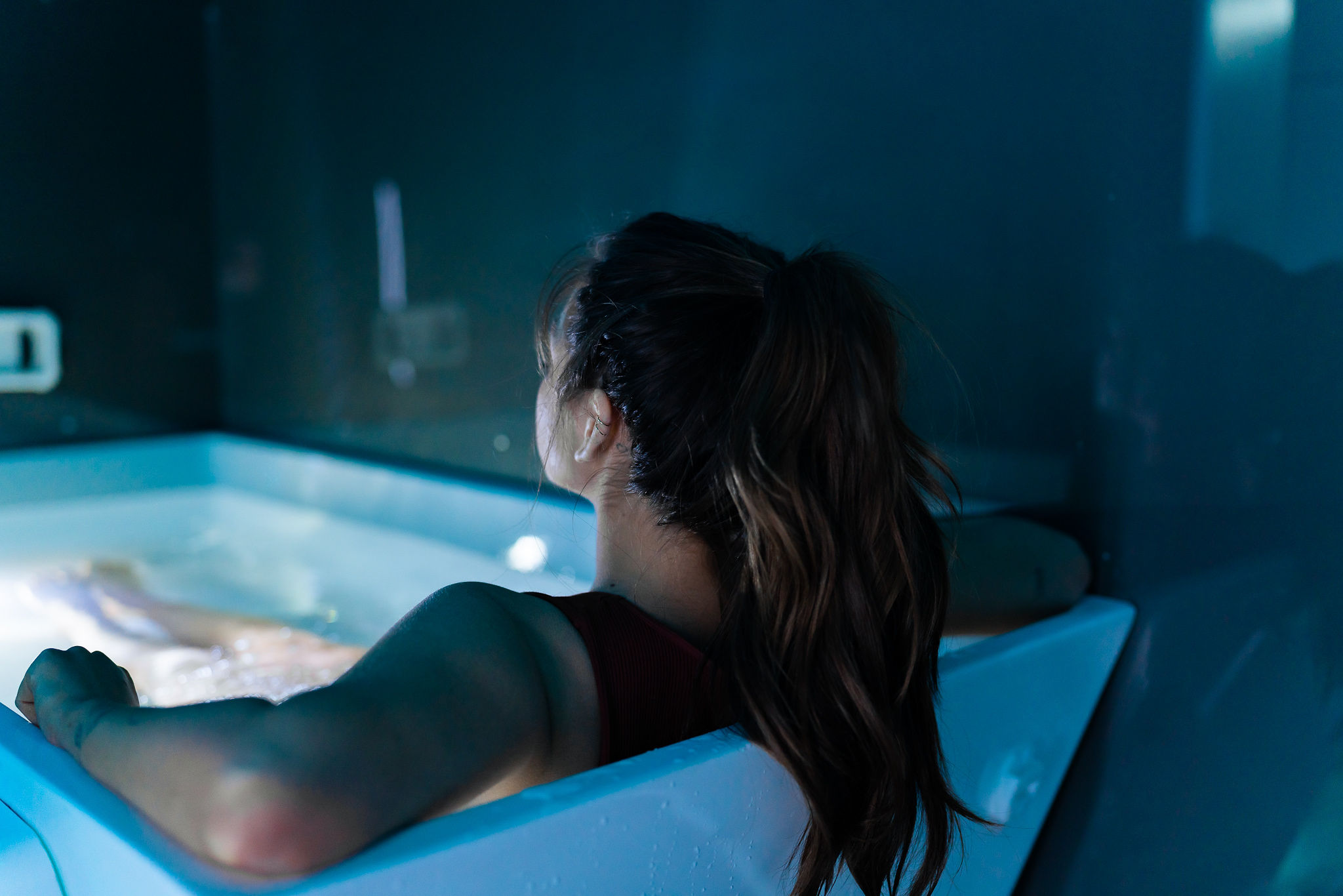 DSC03767 A woman with brown hair in a ponytail relaxes in a white bathtub filled with water, leaning back with her arms on the sides, in a dimly lit room with blue walls.