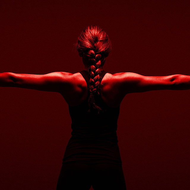 EQXM_EDT_RED_LIGHT_PRIMER_23_243_061523_HERO A person with a braided ponytail stands with arms outstretched, seen from behind, wearing a sleeveless top. The image is lit with dramatic red lighting, highlighting the muscles in their arms and back.