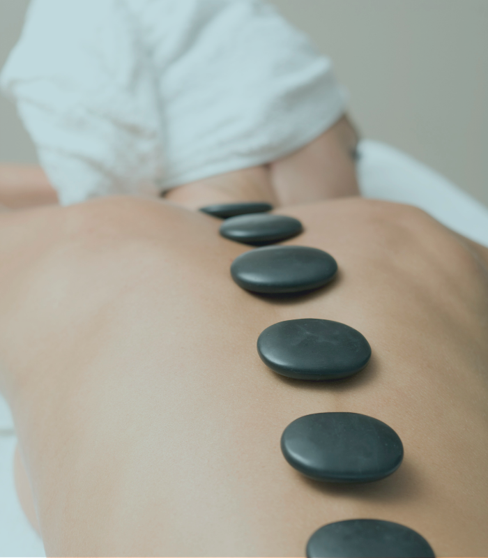 Untitled design (25) A person receiving a hot stone massage, lying face down with a towel covering their head, and several smooth black stones placed along the center of their bare back.