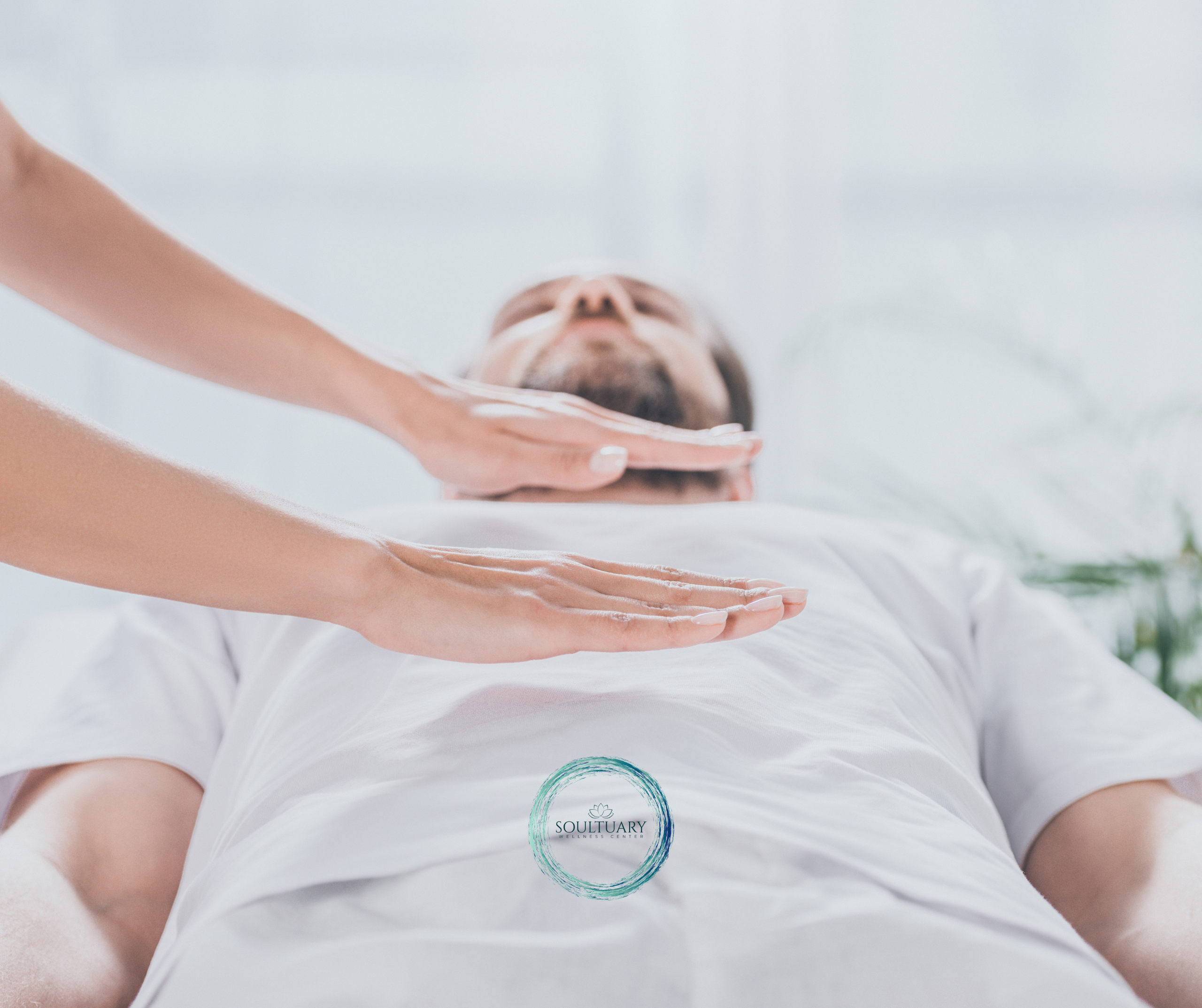 Untitled design (7) A person in a white shirt lies down while another person holds their hands above the persons chest, performing an energy healing or Reiki session. The atmosphere is calm and peaceful.
