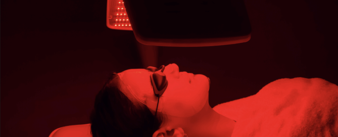 A person lies on a spa bed wearing protective goggles and a facial mask, illuminated by a red LED light therapy device above their face, in a dark room.